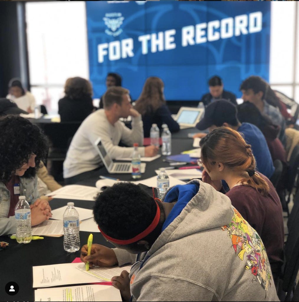 A photo of a long black table where high school students and instructors are writing on paper and laptops and discussing their work. In the background, there is a large window and a blue banner with “For the Record” across it in bold, white block letters. 