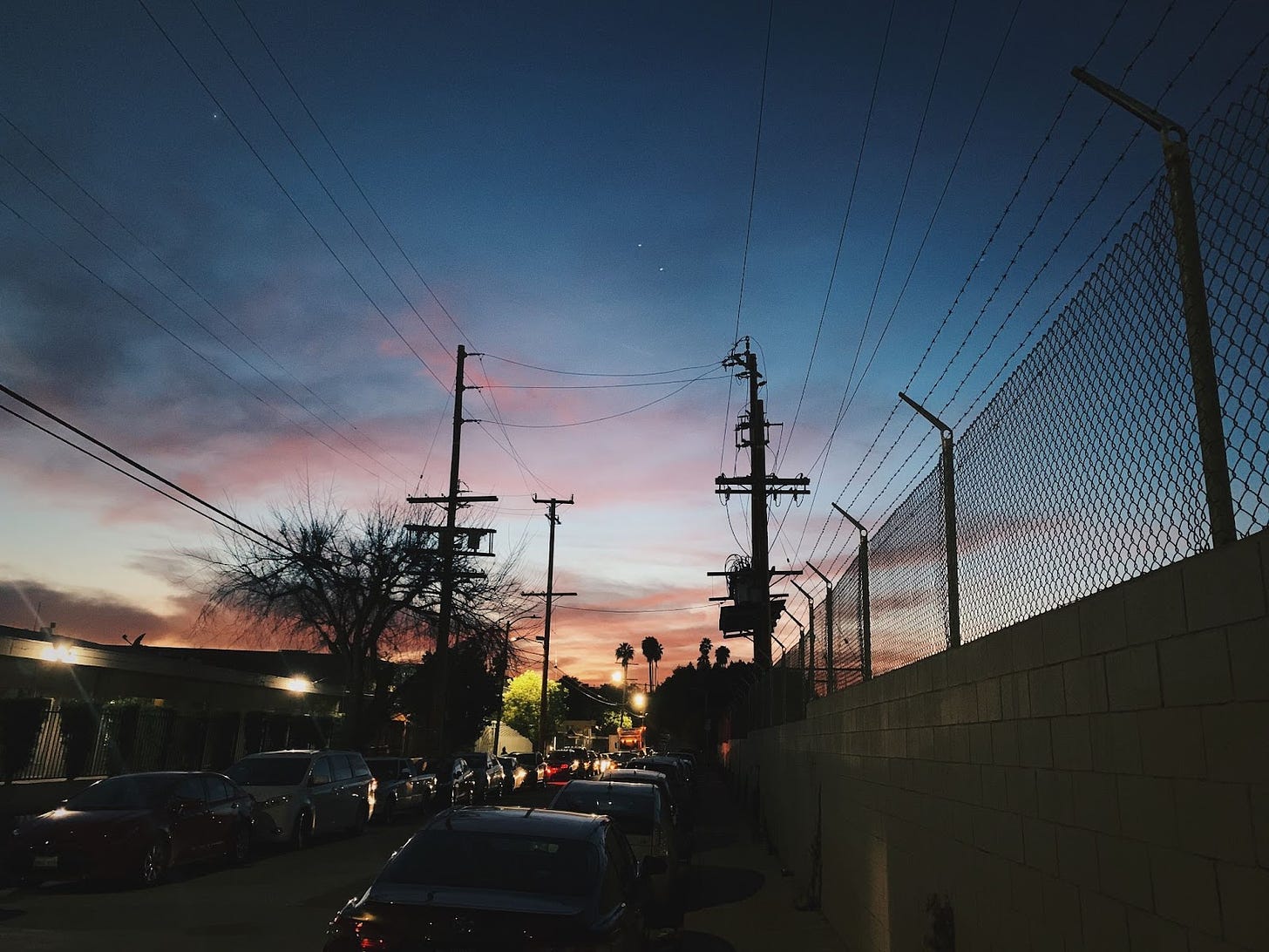 Photo of a city street at dusk with a blue pink orange sunset, power lines, a brick wall with barbed wire, cars parallel parked, street lights, and palm trees at the end of the street.