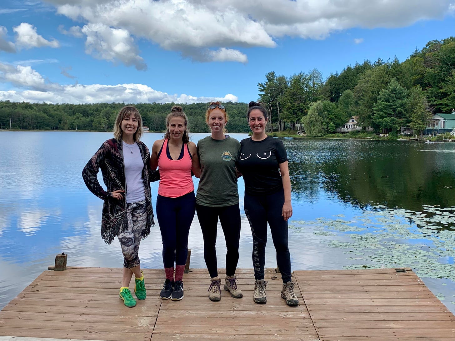 Four women standing on a pier at a lake with trees in the background