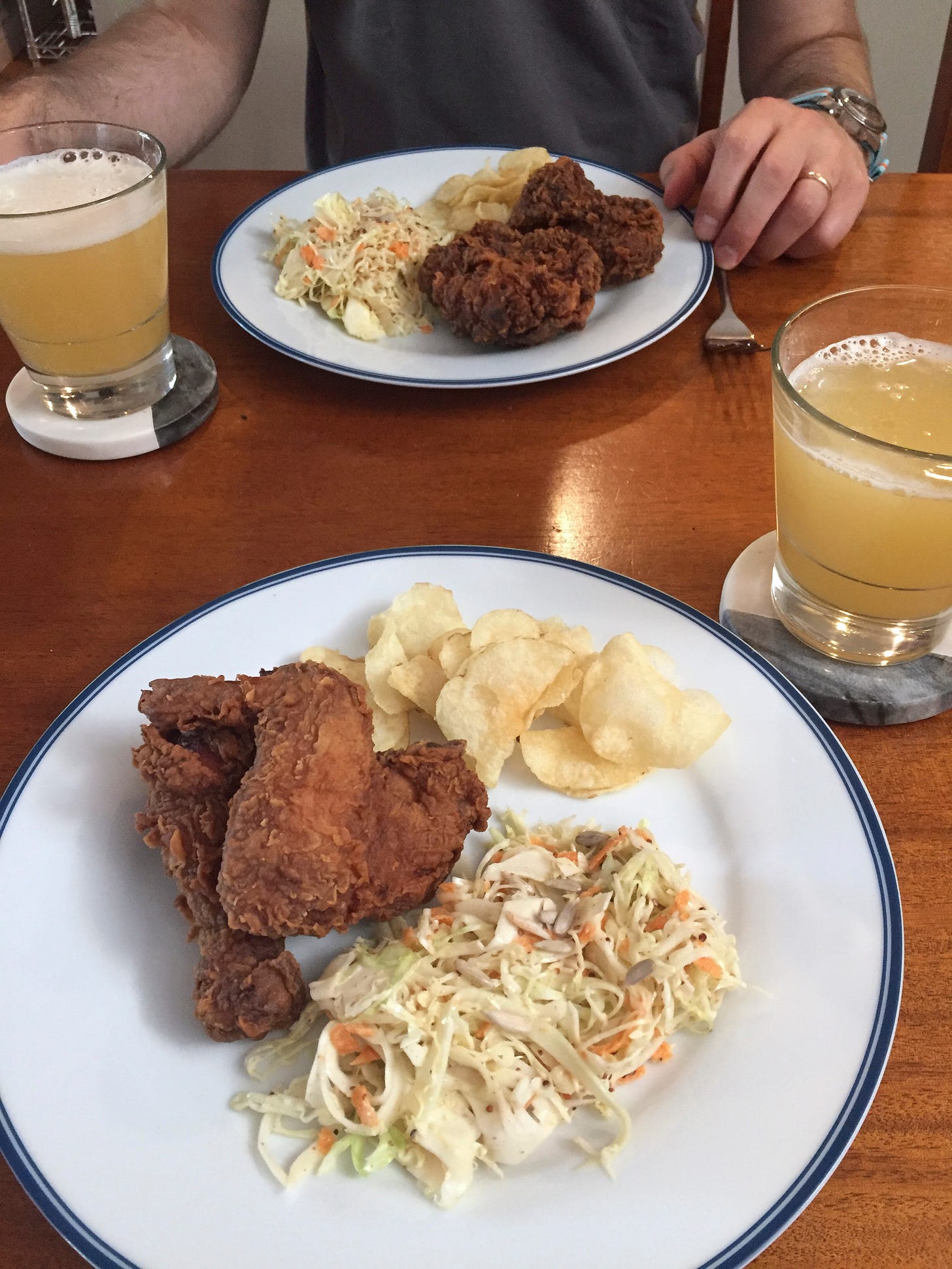 Two white dinner plates, each with two pieces of fried chicken, a small pile of kettle chips, and some coleslaw. Next to the plates are glasses of IPA on coasters.