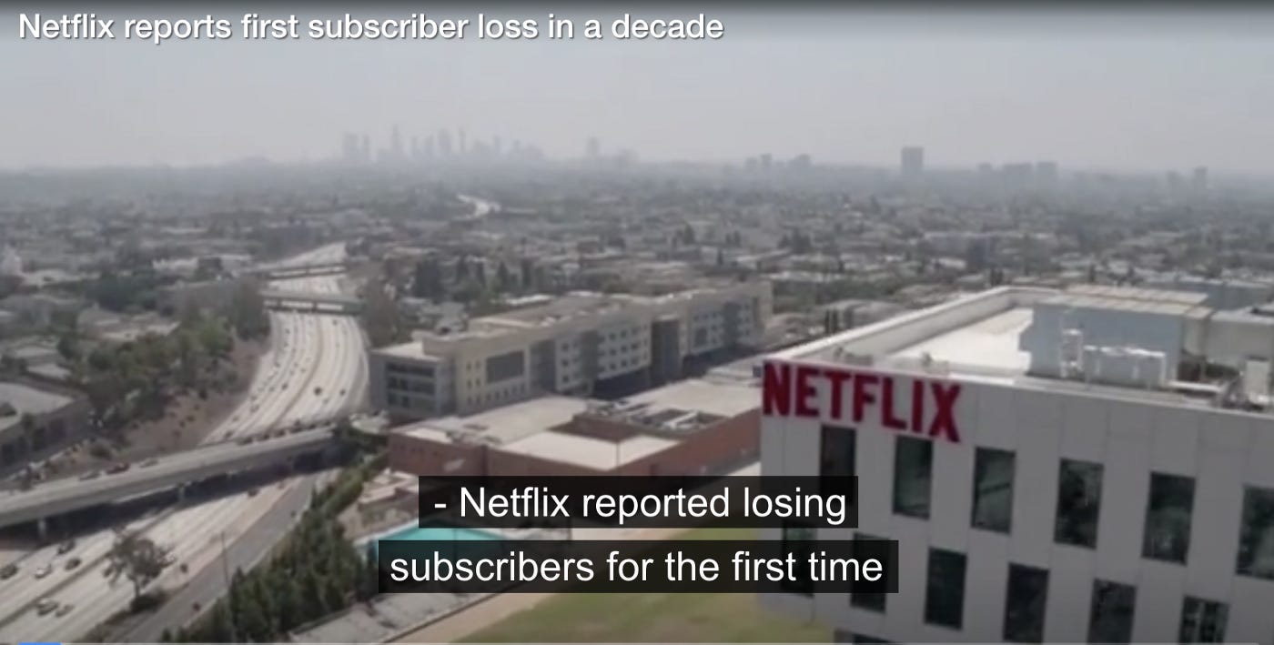 A picture of a city landscape with highways, trees, and office buildings clearly visible in the foreground and skyscrapers visible on the distant horizon that appears hazy with pollution. In the forefront to the right is a building with “Netflix” emblazoned in red on the side. A caption at the top left of the image reads: “Netflix reports first subscriber loss in a decade.” Subtitles at the bottom center read: “- Netflix reported losing subscribers for the first time.”