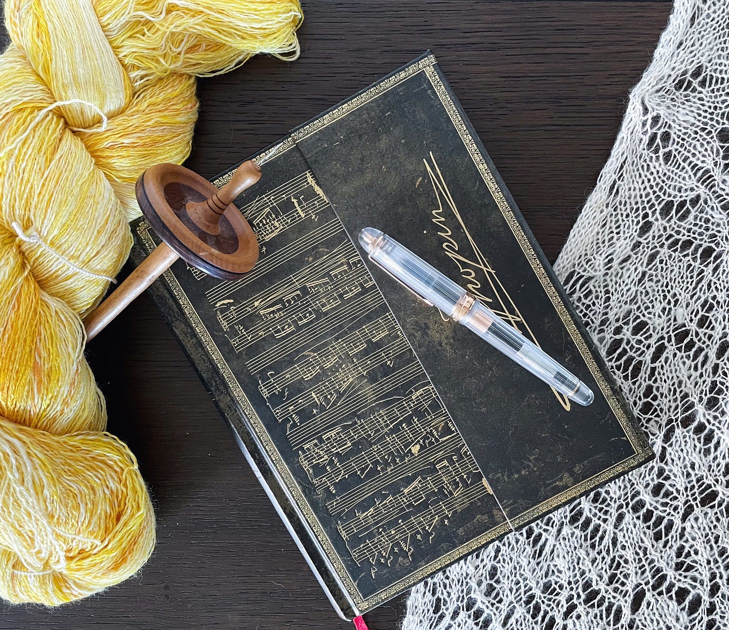 Flatlay of a brown notebook with musical notes on it. Fountain pen on top. A hank of yellow yarn and a spindle to the left. A white lace shawl to the right.