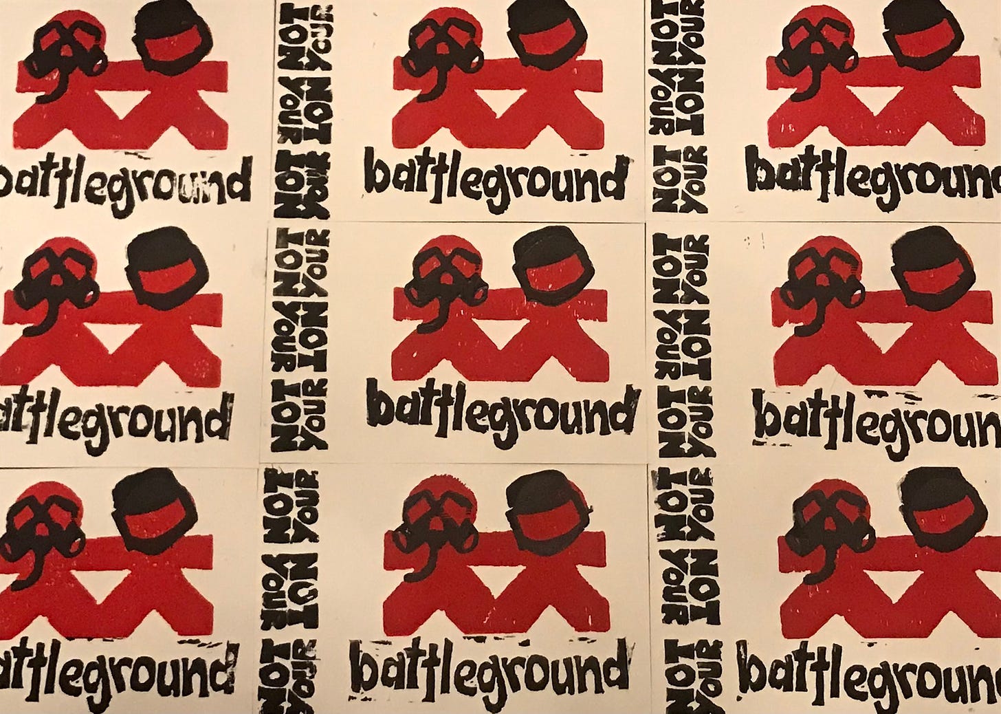 Several prints from an edition of block prints by Anna Caudill. Print features a variation on Vanderbilt Children’s Hospital logo, in which 2 red figures representing children are clad with black bomb helmet & gas mask. Black text along the side and bottom of the figures reads “Not your Not your Not your battleground”.