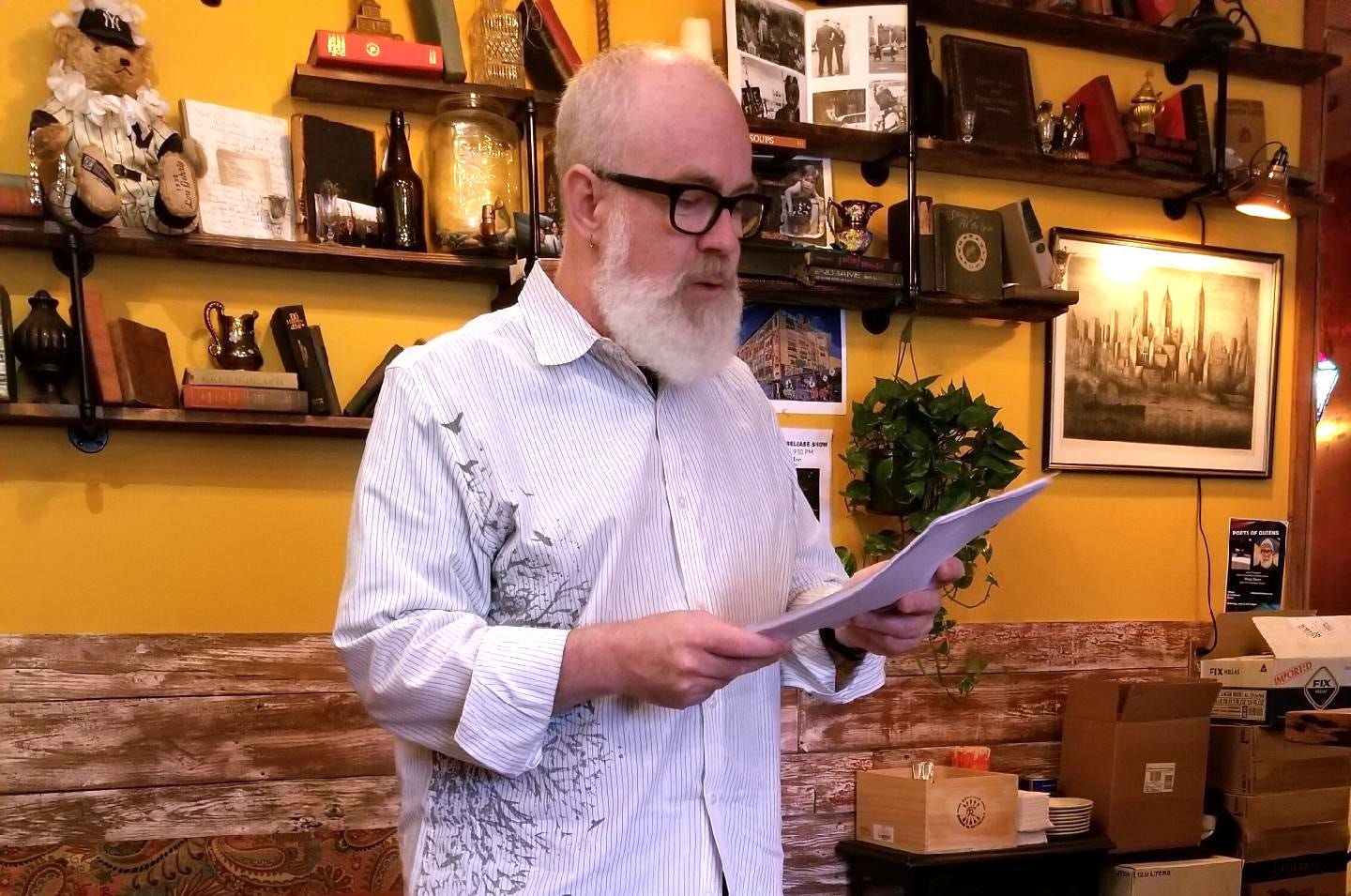 A bearded man in glasses and a striped long-sleeved shirt with a print of birds in a tree reads from a paper manuscript in a warmly lit rustic space before a yellow-painted wall and shelves crowded with books and knickknacks.