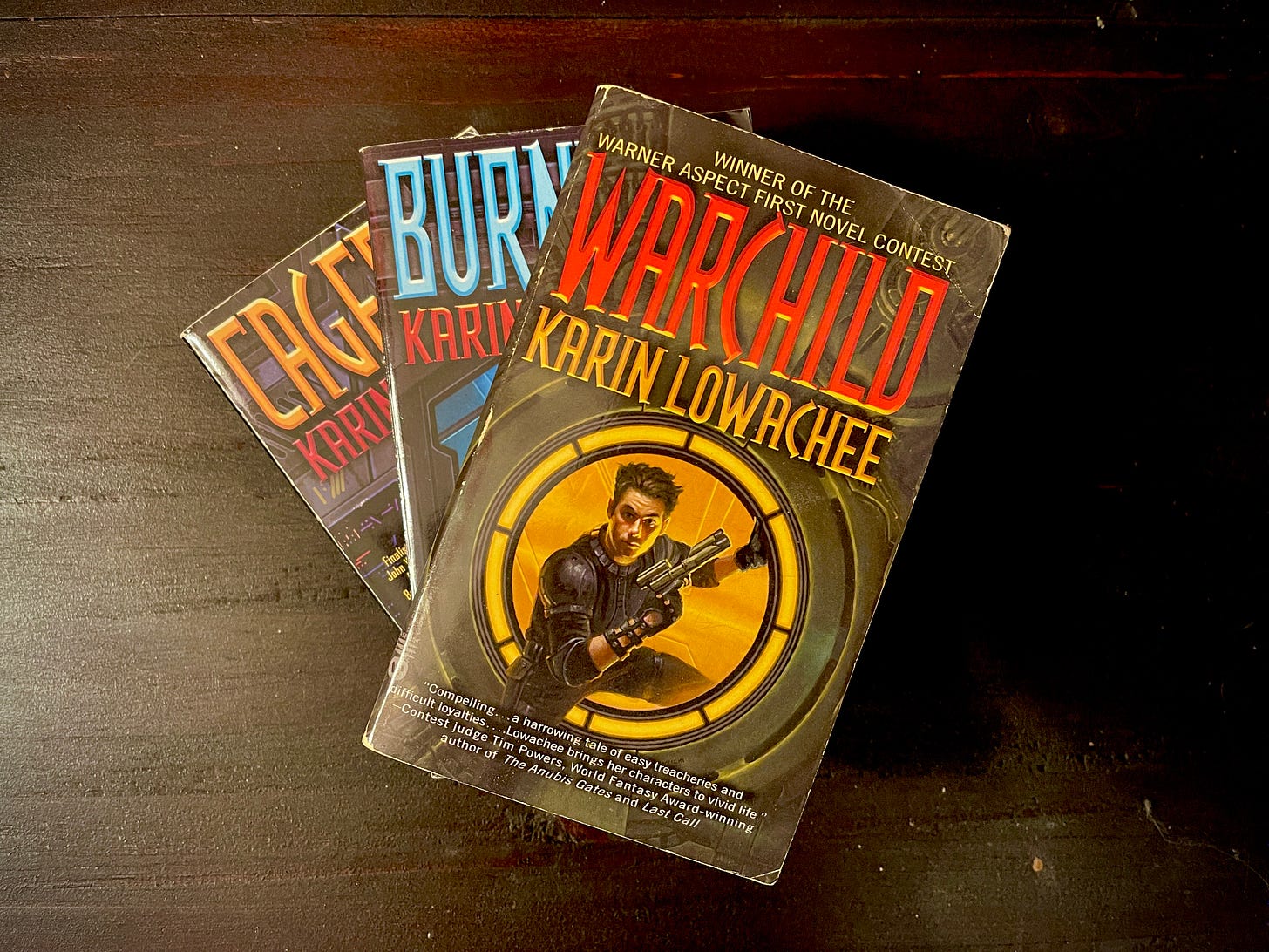 Three books in a spiral stack. The book on the top is Warchild by Karin Lowachee, and features a person on the cover stepping through a portal with a gun. 