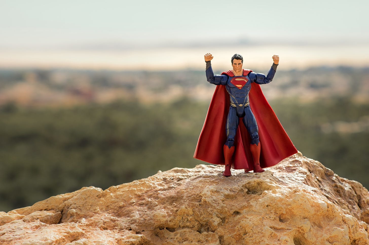 A small Superman toy rests on a rock with its arms raised in the air.