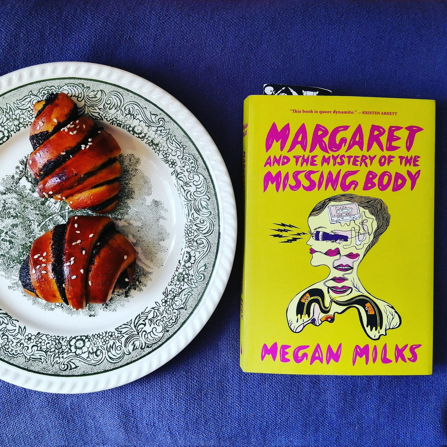 Auf einer blauen Decke liegt das Buch "Margaret and the mystery of the missing body" von Megan Milks, daneben steht ein weiß-grün gemusterter Teller, auf dem zwei Gebäckstücke liegen 