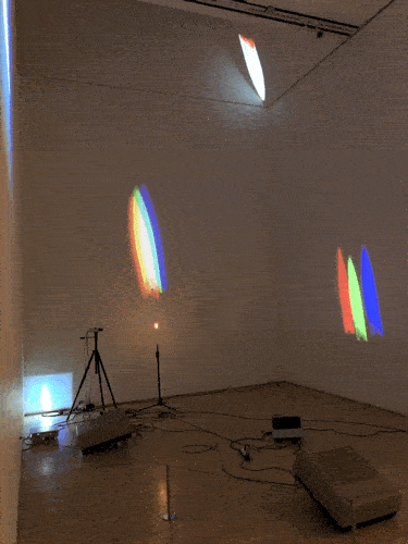 “One Candle (Candle Projection)” by Nam June Paik, 1989.  Closed-circuit television (CCTV) camera, tripod, lit candle on custom stand, video projectors, and modified cathode-ray tube projectors