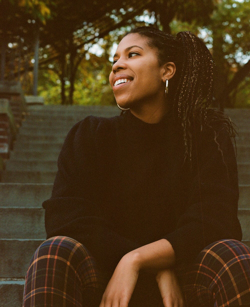 Nia, a Black person with long hair, in a black sweater and plaid orange and blue and black pants - sitting on stone steps looking to the left and smiling coyly