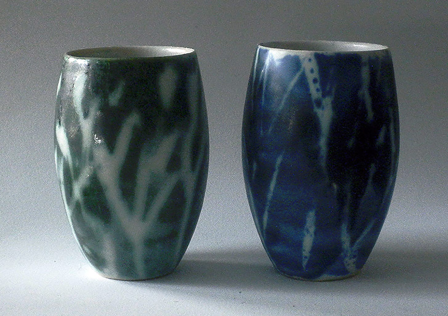 Two slip cast Tumbler are simple forms of gently curved ceramic drinking vessels glazed in white and then brushed with wax branches and brushed again with with fluid interactive ceramic decorating color. One tumbler is brushed in green and the other in blue, The interaction between the flux in the decorating colors and the white glaze created a nature-made effect