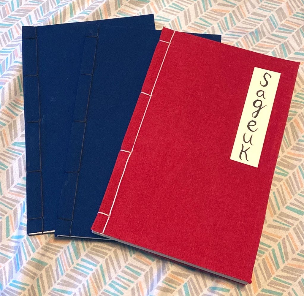 Three books bound in the traditional side-stitched style. Two have dark blue covers with brown binding thread. The one on top is in red with white thread and has a name-plate which reads "Sageuk"