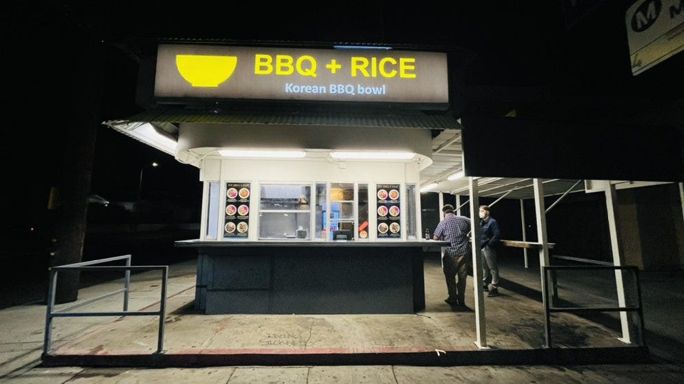 A photo taken at night of the shack after its conversion to BBQ + Rice. The shack is now grey on the top and bottom, with white and windows in the middle. The sign says the business name in yellow. Two men stand outside the business which is otherwise deserted. 