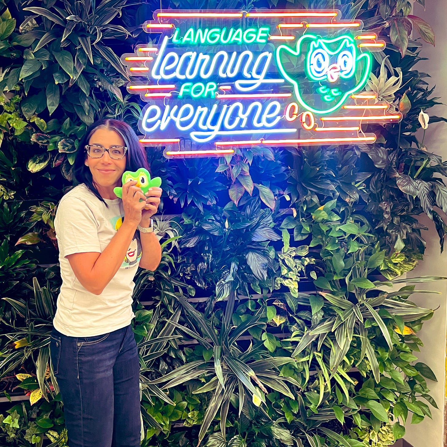 Photo of me in blue jeans and a cream t-shirt with Sally Wiggin popart on the front standing in front of a wall of greenery while holding a small stuffed green Duo owl. There is a neon sign on the greenery wall that says "Language learning for everyone."