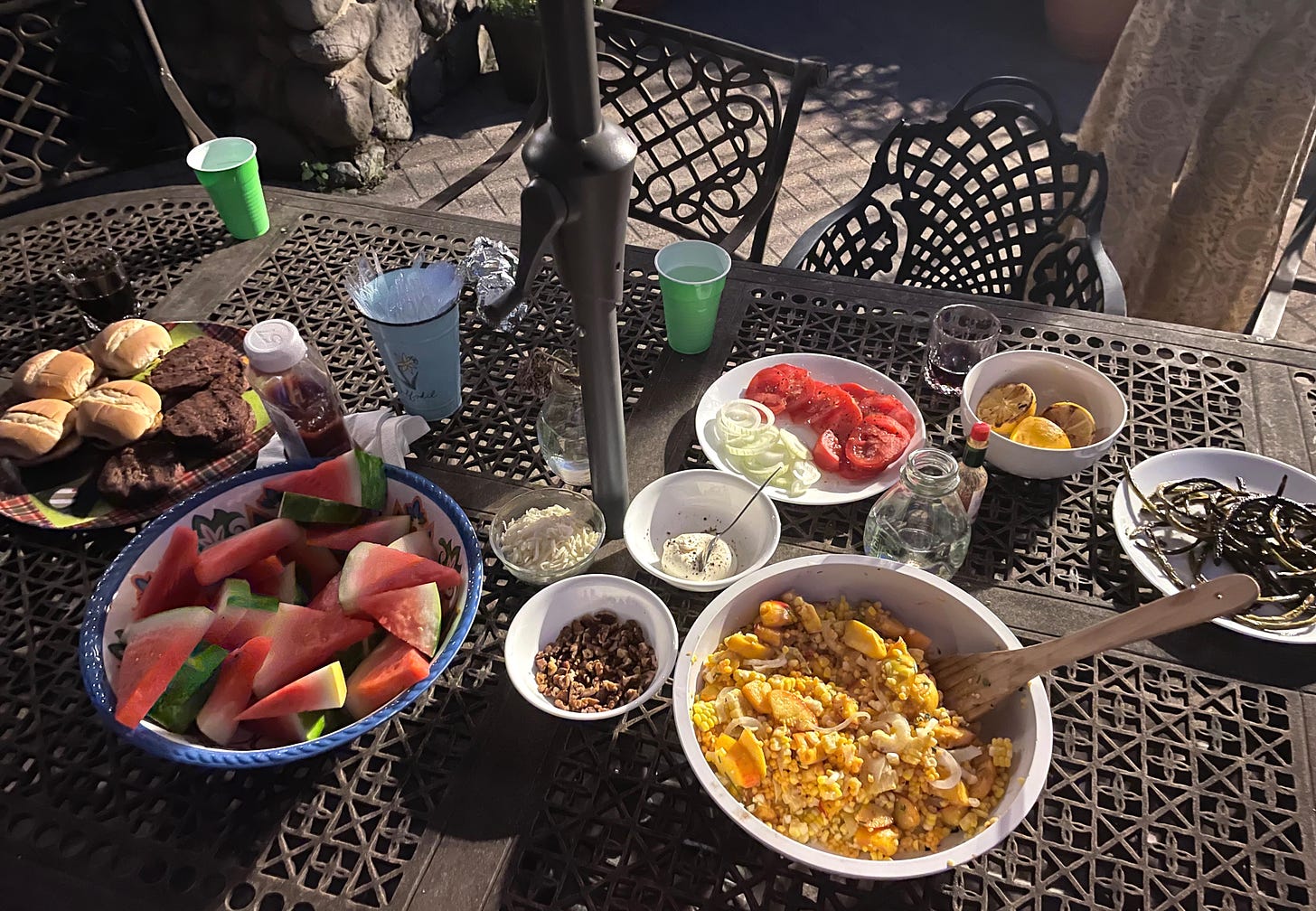table with impossible burgers, watermelon, peach and corn salad, garlic scapes, grilled lemons, and sides of toasted walnuts, burrata, and tomato and onion slices