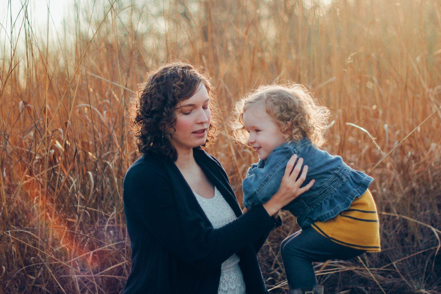 Adult with brown curly hair lifts a smiling toddler with blonde curly hair, in a wheat field