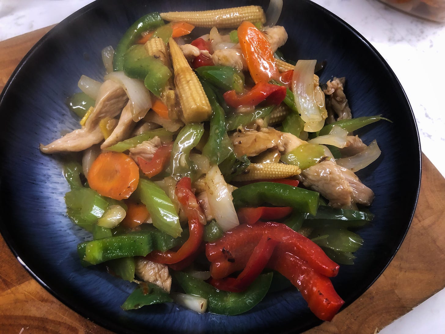 A large bowl filled with stir-fried chicken and vegetables including red and green pepper, onion, carrot, baby corn, celery, and green onion