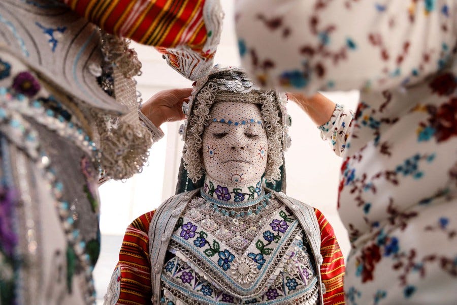 A bride, her face covered in white makeup lined with patterns, has two people help with her traditional clothing.