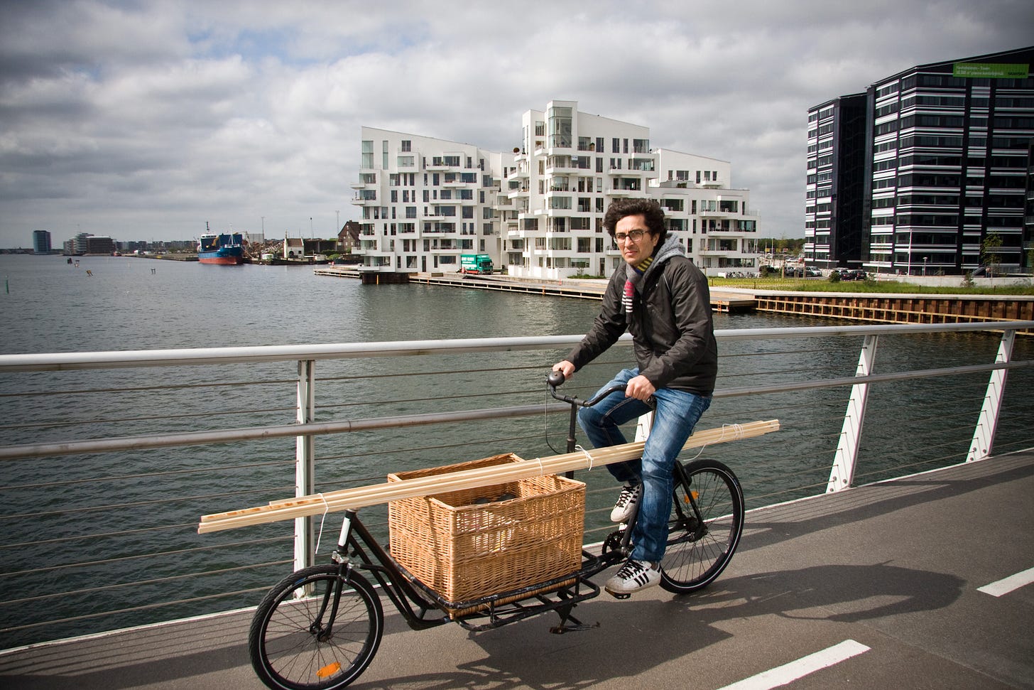 A cargo bicycle passes over a bridge in Copenhagen. In the background, new apartment buildings stand next to the water. The cargo bicycle is carrying a thin plank of wood.