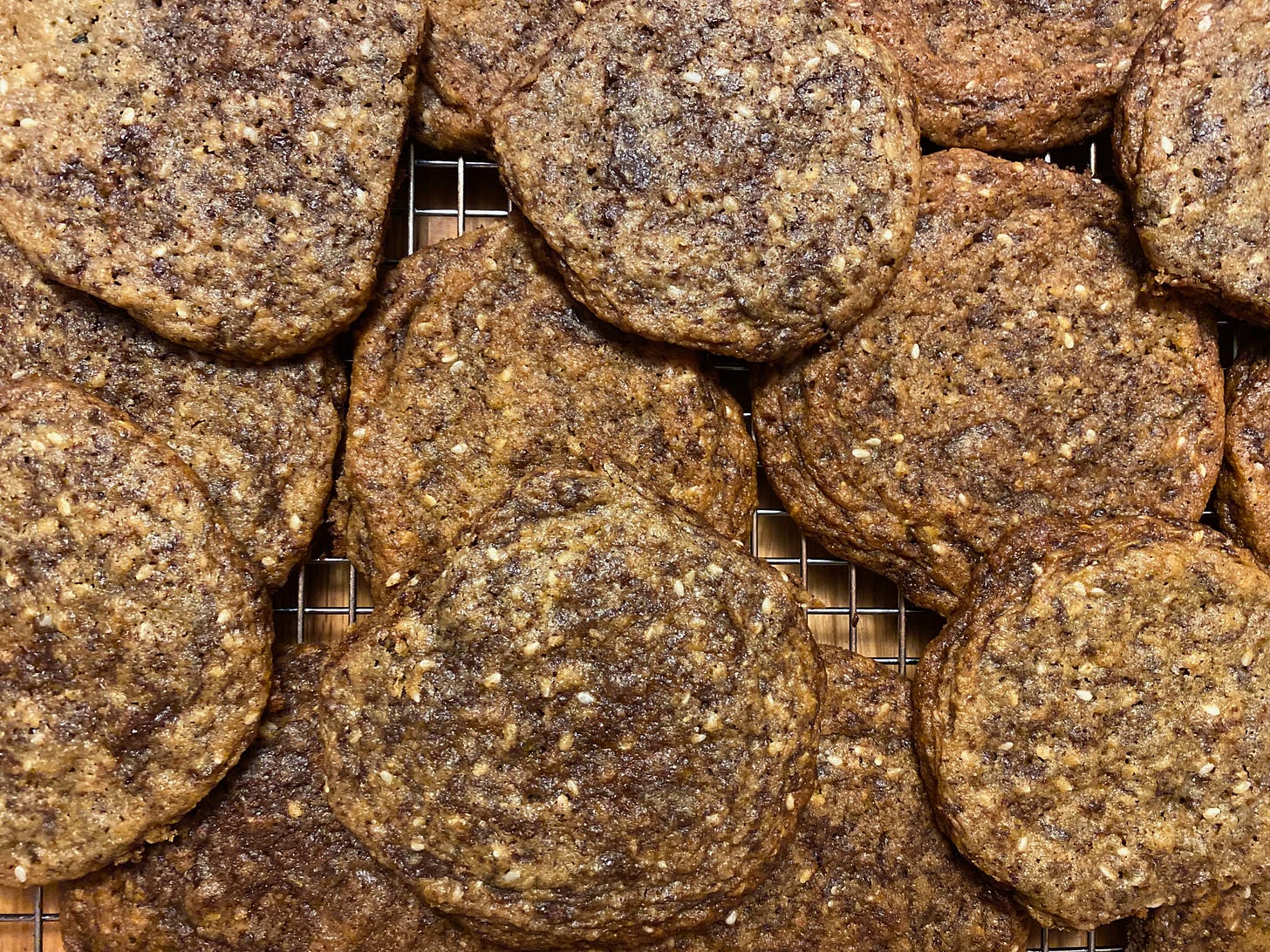 A close-up of a pile of round, crinkly chocolate chip cookies on a metal cooling rack. They are golden brown and studded with sesame seeds and chunks of chocolate.