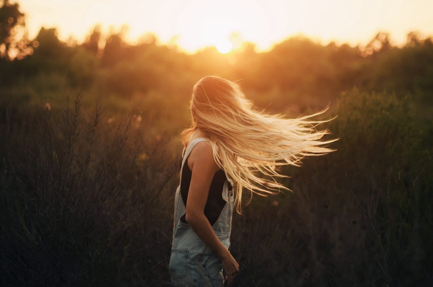 A woman with long blond hair dances in a field at sunset.
