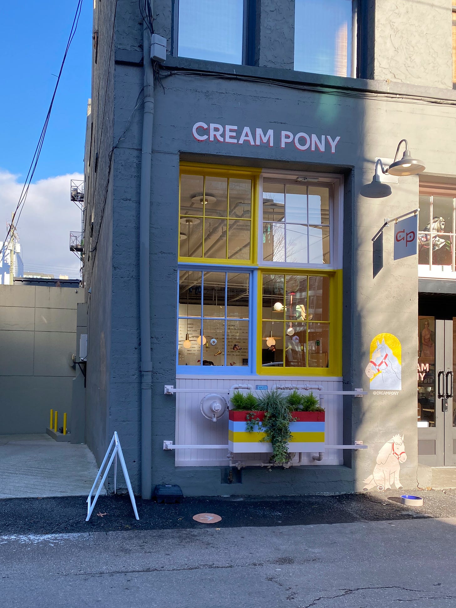 A grey building with the words 'Cream Pony' in pink and red block letters. Below the words are four panels of windows, their frames painted yellow, blue, and pink. A striped planter box is below them with plants trailing out of it. Next to the window are two paintings of horses, below a sign labelled 'CP'. Behind the building, a fluffy white cloud sits low on the horizon, and the sky is blue.