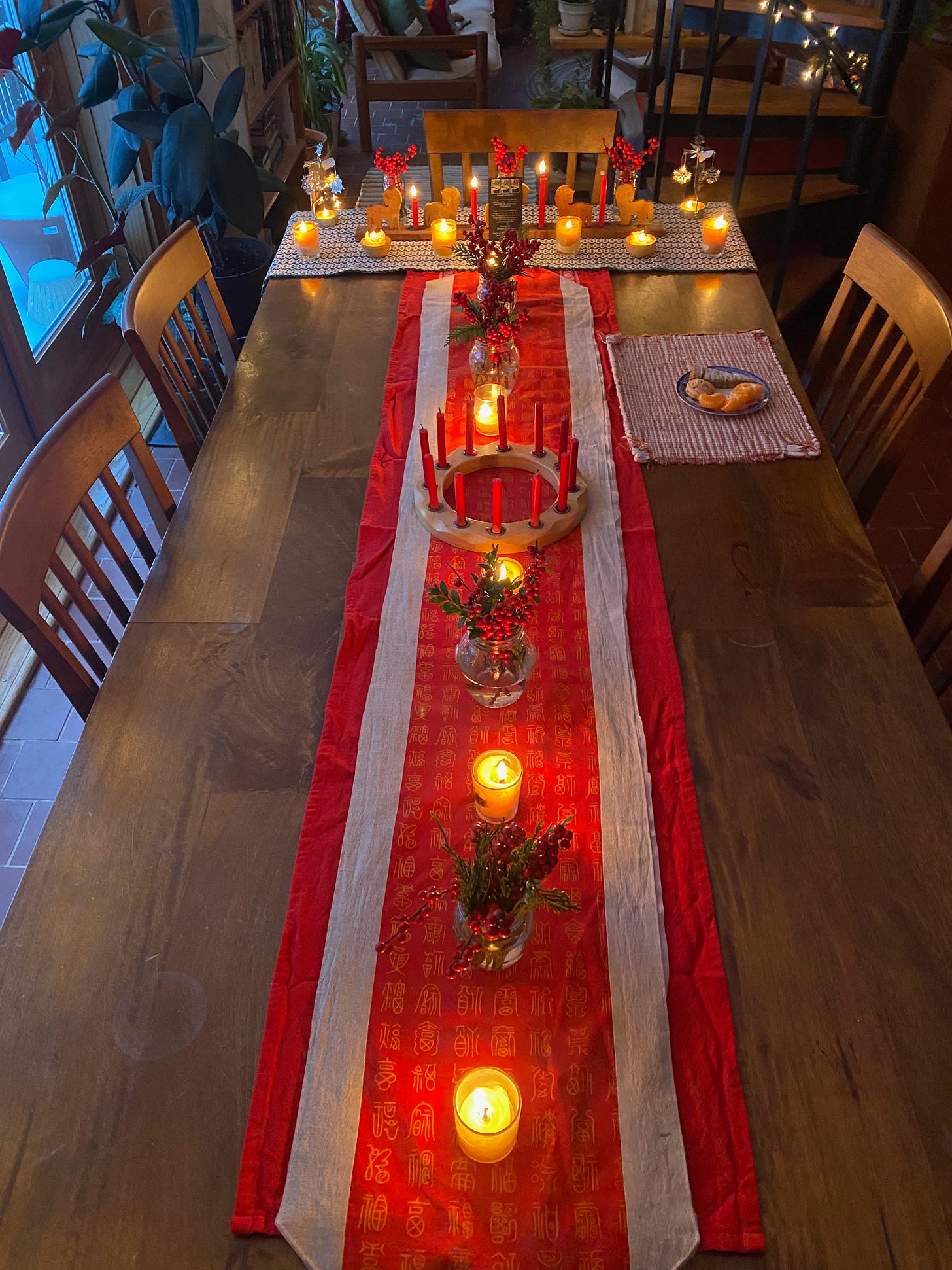 View from above of a table with red runner. Arranged on the runner are alternating jars of winterberry and lit candles. A circular candleholder sits in the middle, and at the end of the table is a green and white woven cloth with more candles and jars of winterberry.