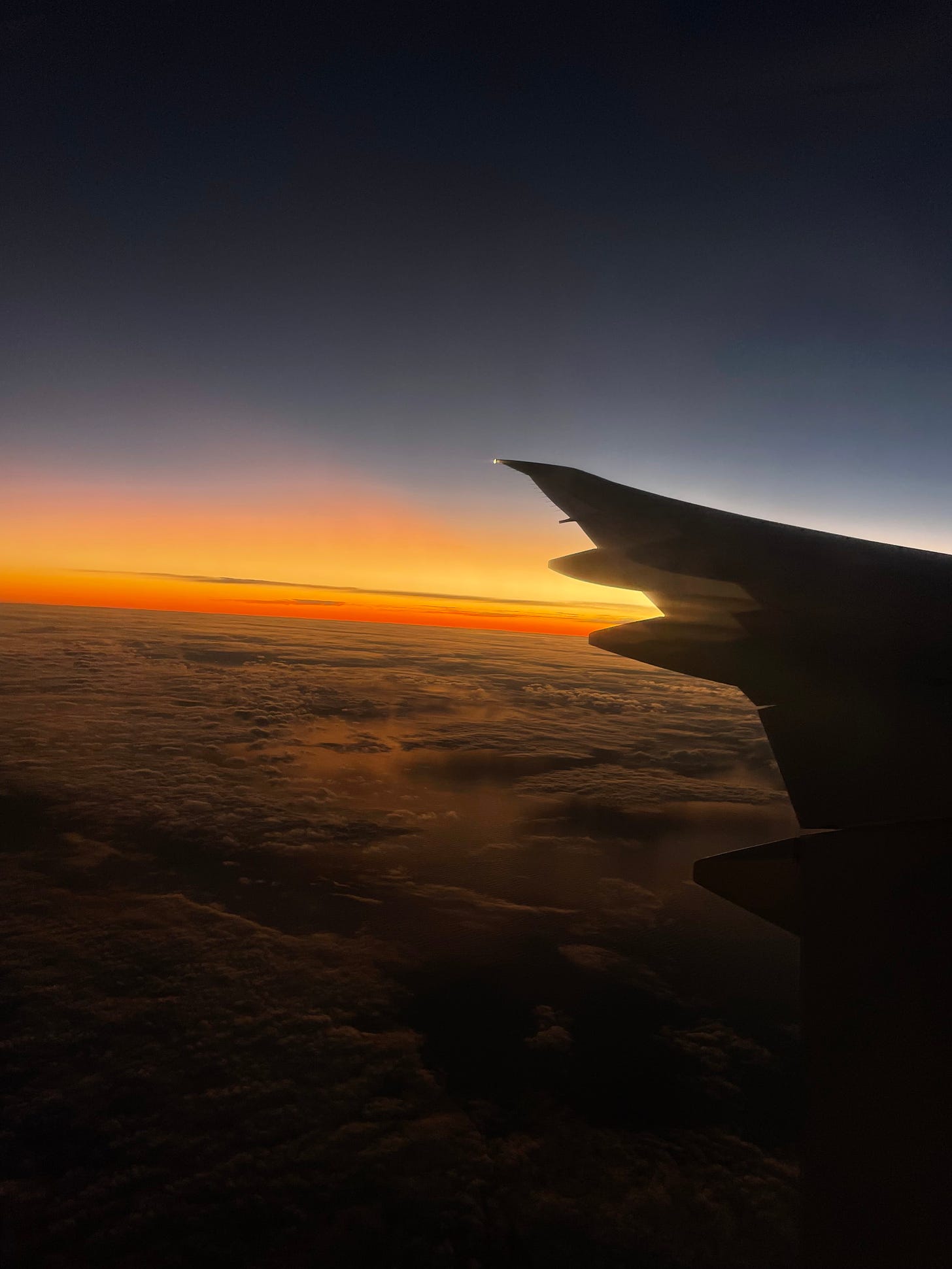 View of sunrise from a plane window. Sharp oranges and soft yellows blend into the deep blue sky. Clouds drift over the Pacific Ocean below. The silhouette of a plane wing.