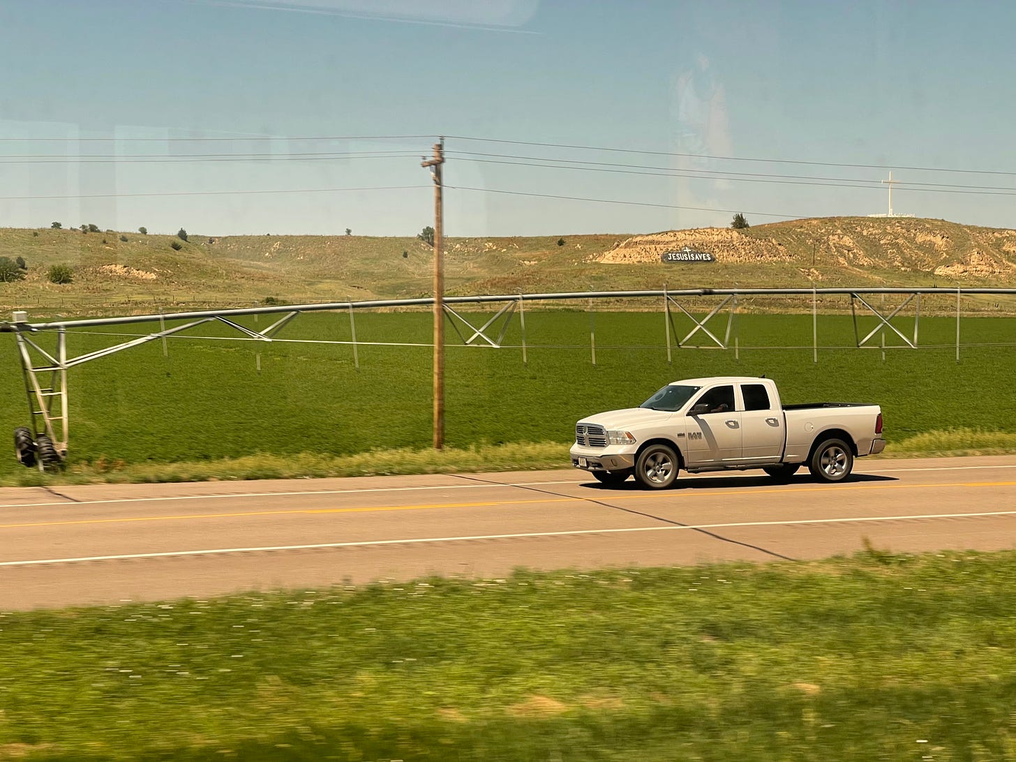 a highway in rural Colorado, with a white Ram truck. an irrigation boom spans a planted field. “Jesus Saves” is painted in large white letters on a hill in the background.