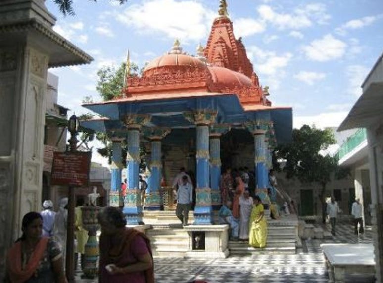 A picture of the outside of a building during the day. The floor is white and black marble, with white marble steps ascending into the temple. There are people in traditional dress on the steps and walking around the floor. The pillars of the building are sky blue with carvings painted light pink and green. The carved domes are saffron colored with golden tops.