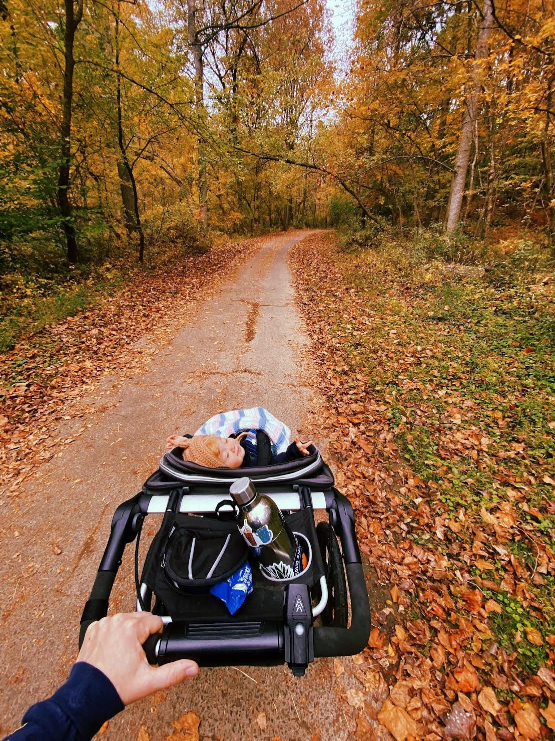 paved path cutting through woods of fall foliage, black stroller with baby looking up, closed eyes at runner who's hand is on pushing the stroller