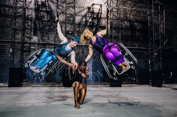 A “Wired” rehearsal picture of Laurel Lawson, left, and Alice Sheppard suspended in the air in their wheelchairs at a 90-degree angle, their heads almost touching. Each has an arm holding onto Jerron Herman, who is kneeling on the ground as an anchor. Lawson is a white dancer with blue dyed hair; Sheppard is a multiracial woman; Herman is a Black man with close-cropped hair and barbed wire circling his arms, one of which reaches up to rest on Lawson’s lap.