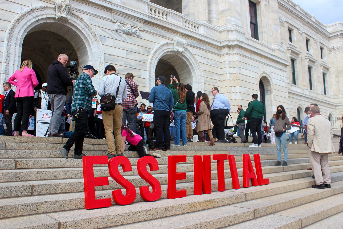 a crowd gathers on top of the capitol steps in front of big red letters that spell out "essential"