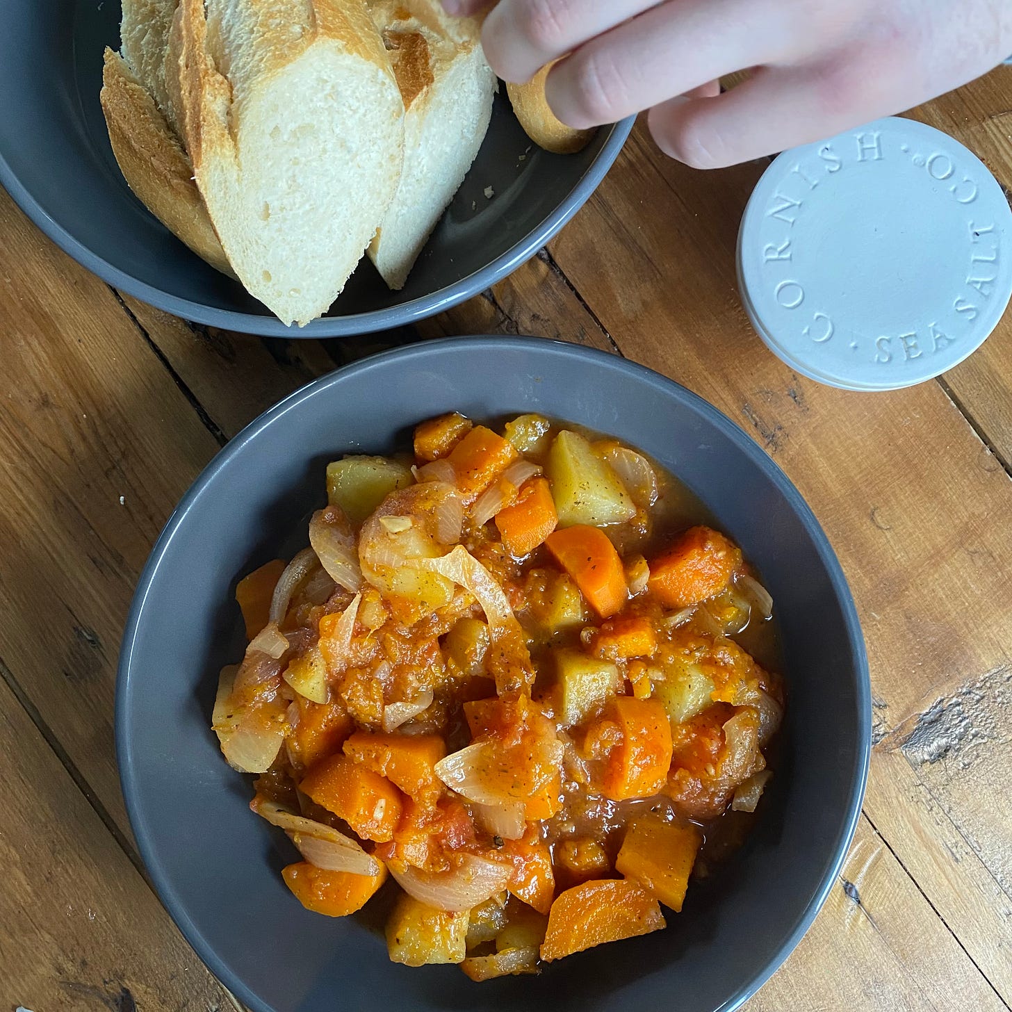 Bowl of spiced vegetable stew in a grey bowl, with a hand reaching for a piece of bread