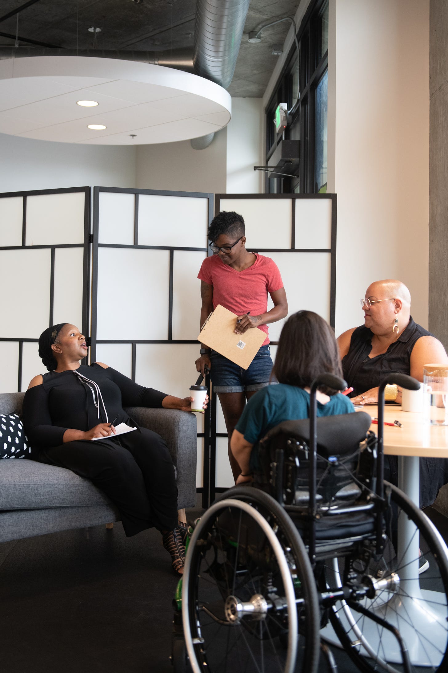 four disabled people of color gather around a table during a meeting. A Black woman sitting on a couch gestures and speaks while the three others (a South Asian person sitting in a wheelchair, a Black non-binary person sitting in a chair, and a Black non-binary person standing with a clipboard and cane) face her and listen. Image Credit: Chona Kasinger via Disabled and Here