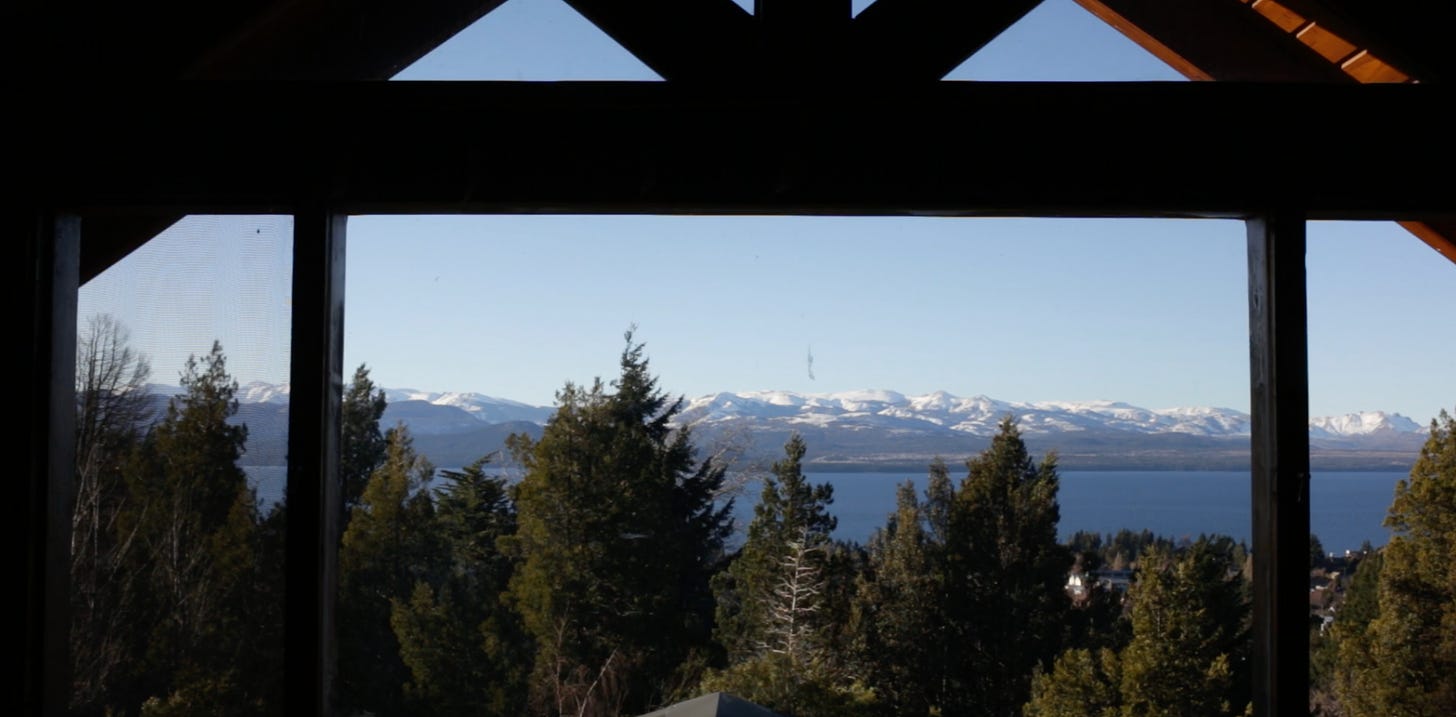A view outside a window in Argentina. In the near view, there are evergreen trees, and as we look farther out to the horizon, there is a blue lake bordering snow-peaked mountains in the distance. It's a sunny, cloud-less sky day.