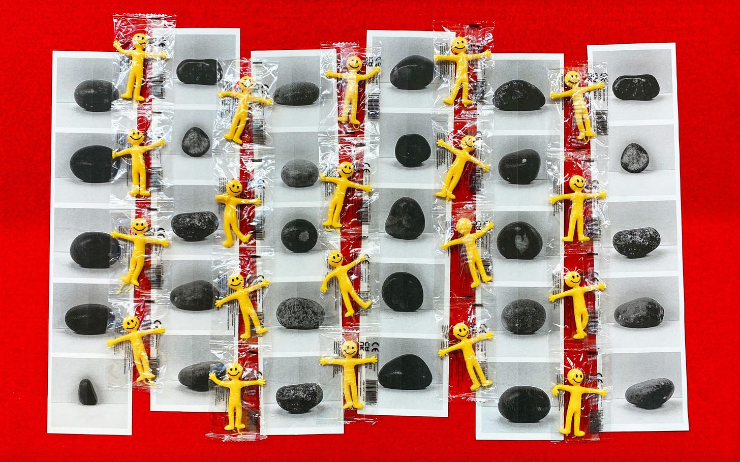 An illustration by Kaiya Waerea features a grid of black and white photos of stones, interspersed with yellow smiling rubber toy figures wrapped in plastic.