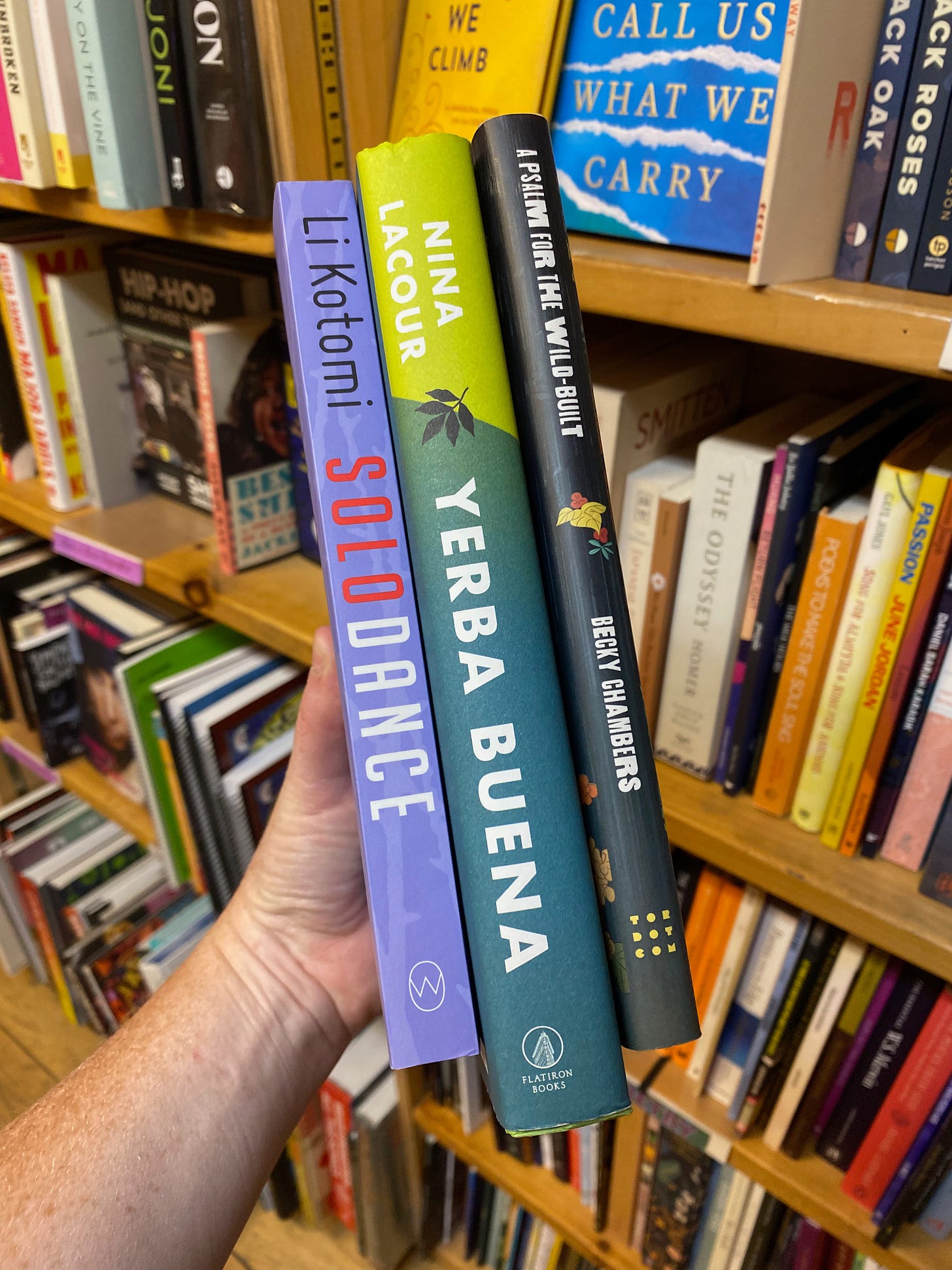 My hand holding these three books in front of a shelf of poetry in a bookstore.