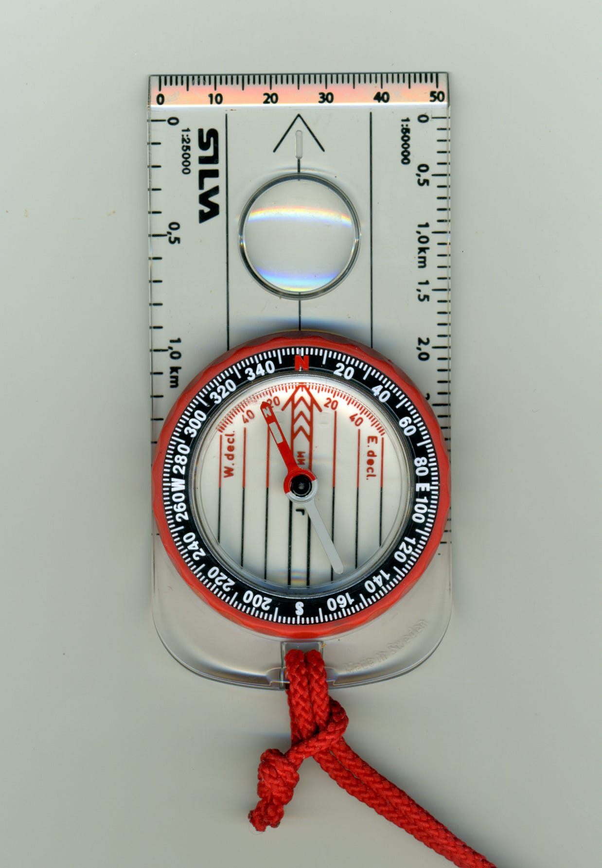 A picture of a compass; a orange cord is tied to one end. The dial and compass arrow are a reddish orange color. The compass is transparent but has markings and rulers on the sides.