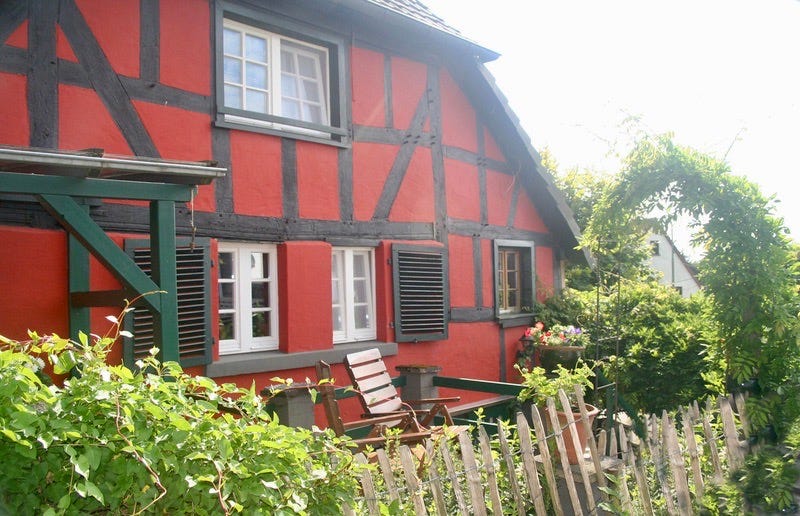 A bright red painted half-timbered house behind a rickety fence, with lots of bright green foliage around