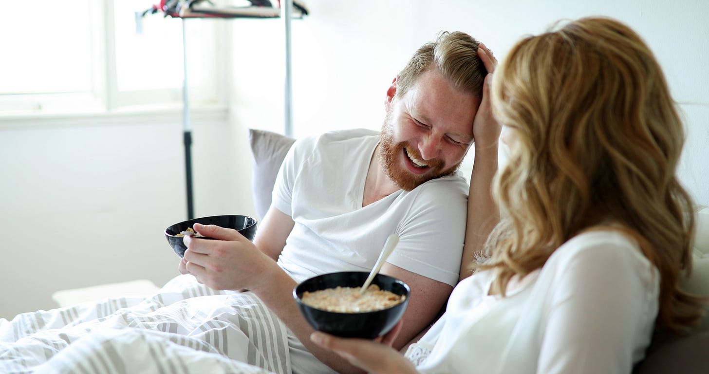An attractive young couple laughs as they enjoy breakfast in bed.