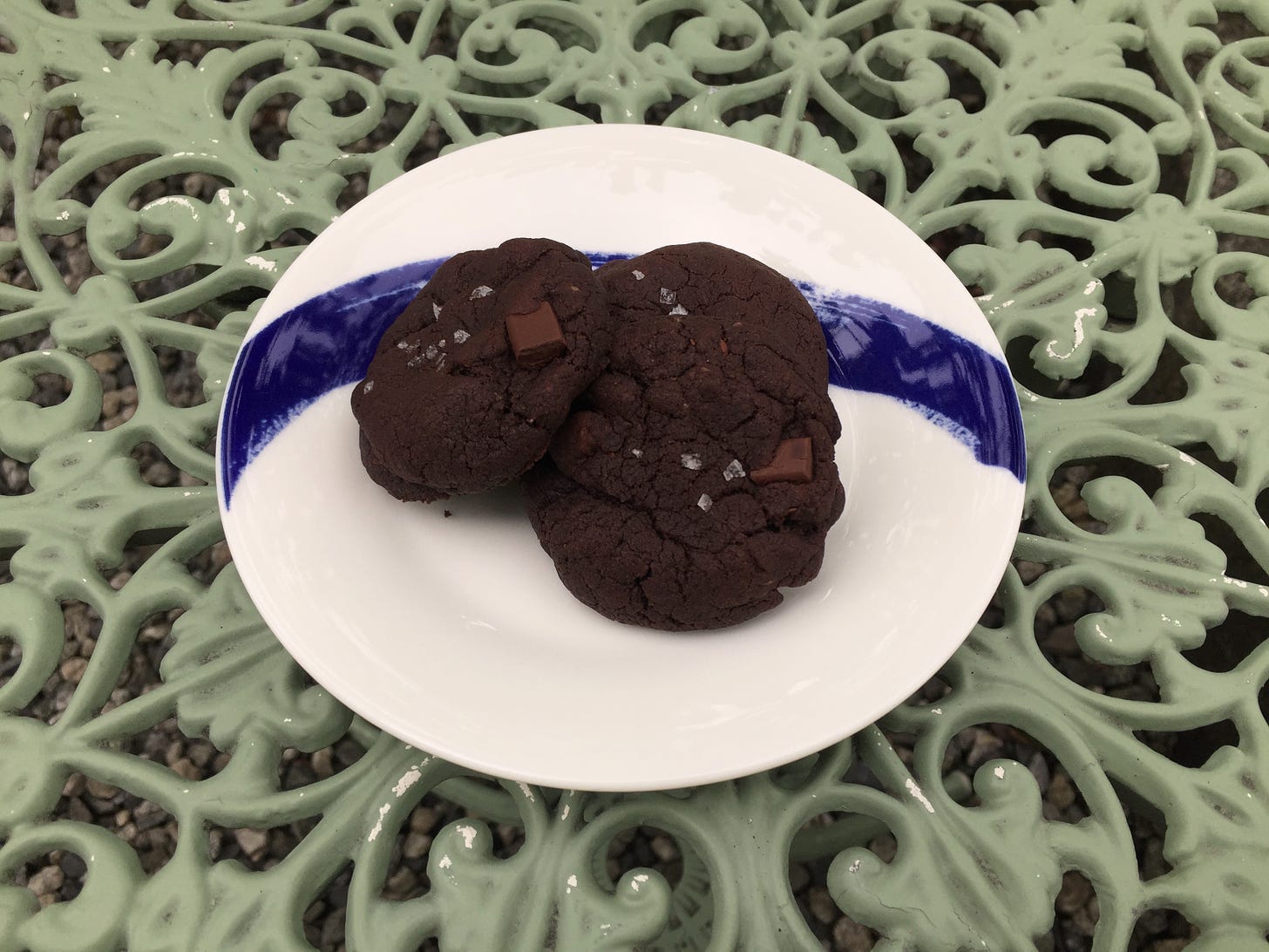 On a mint green wrought-iron table is a small white plate with a blue stripe, which holds three double chocolate cookies with flakes of salt on the tops.