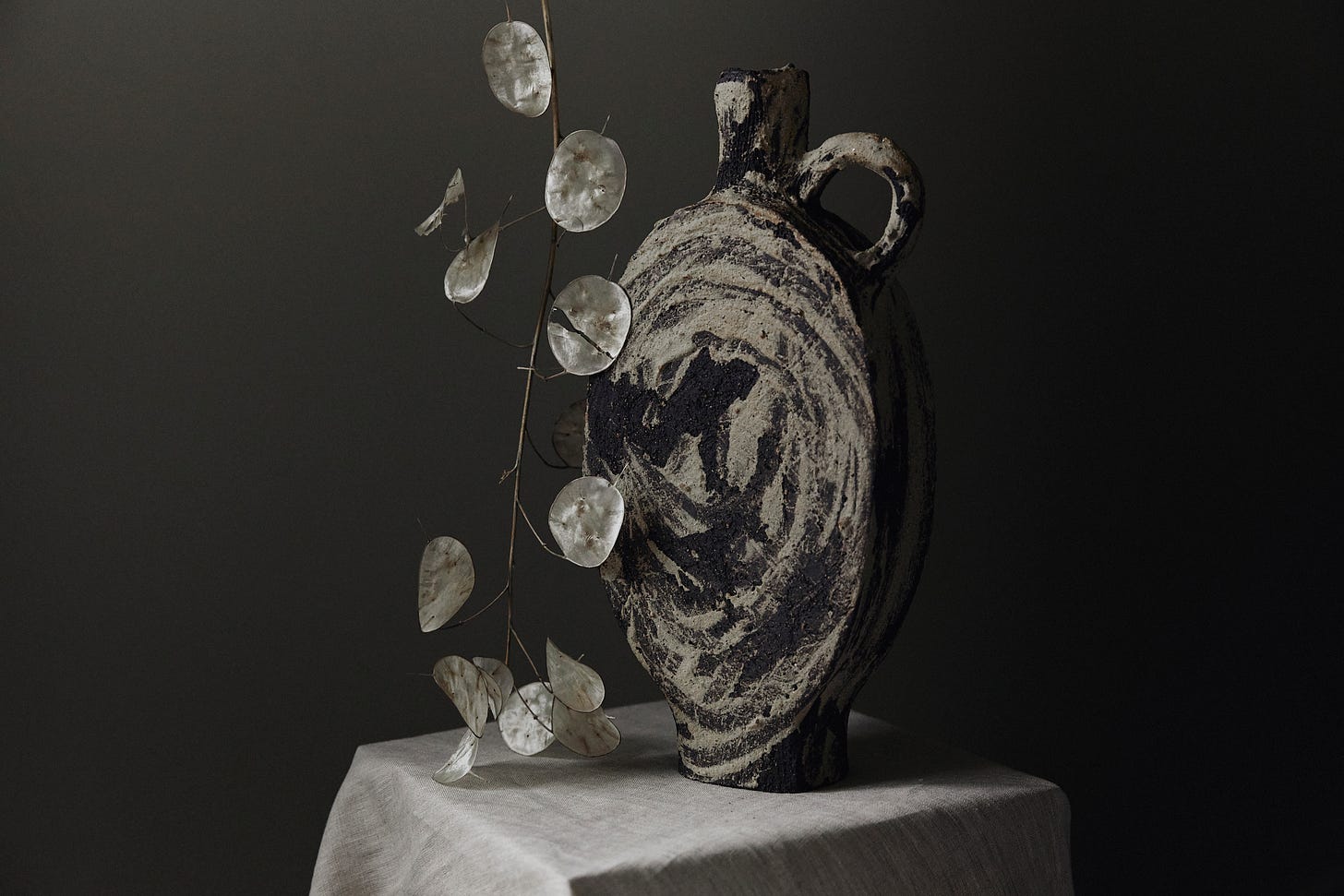 A tall ceramic vessel on a fabric-covered plinth. A stem of winter honesty leans against the vase.