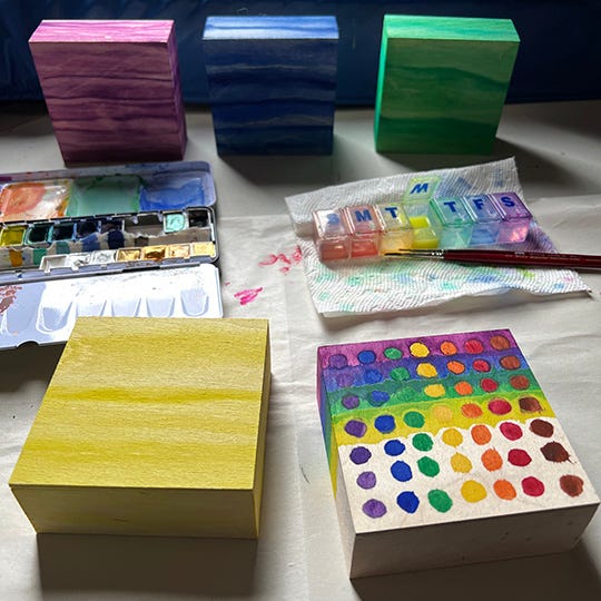 A still photo showing the painting of a 4 inch by 4 inch wooden canvas each week in a solid color, along with another canvas painted with seven rows of seven circles, one each of purple, blue, green, yellow, orange, red, and brown. This shows four weeks into the counting of the Omer, with a purple, blue, green, and yellow canvas and as many stripes on that grid.