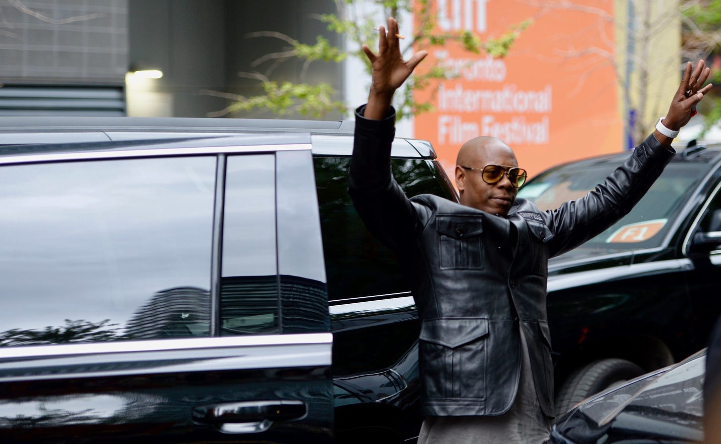 Dave Chappelle holding up his arms in greeting outside a limo