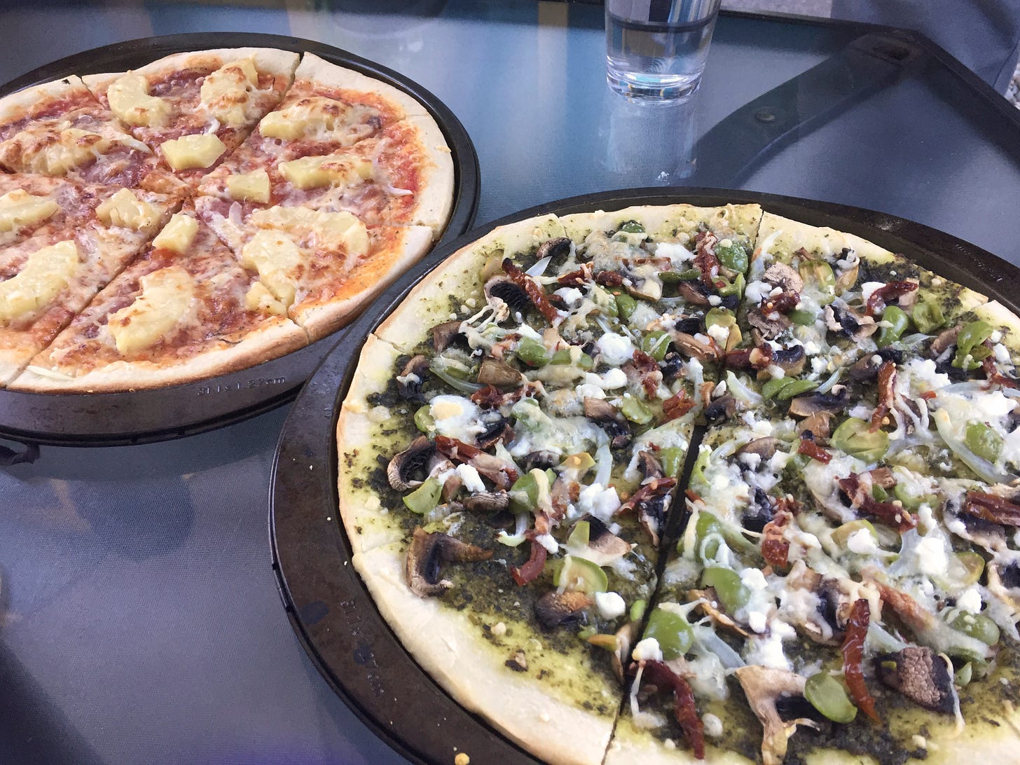 Two pizzas in their pans. In the background, a red sauce pizza with salami and pineapple. In the foreground, a pesto base with olives, mushrooms, sun-dried tomatoes, and feta.