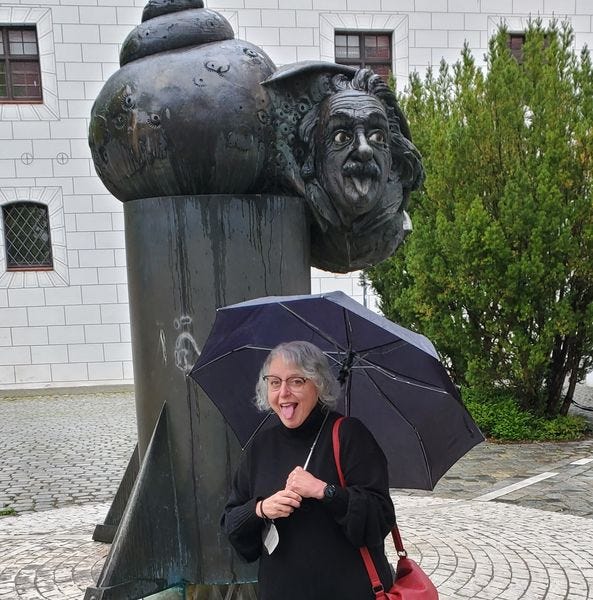 A black stone fountain with Albert Einstein's head on a snails body, sticking out it's tongue, and an older white woman dressed in black holding an umbrella and imitating his expression. 