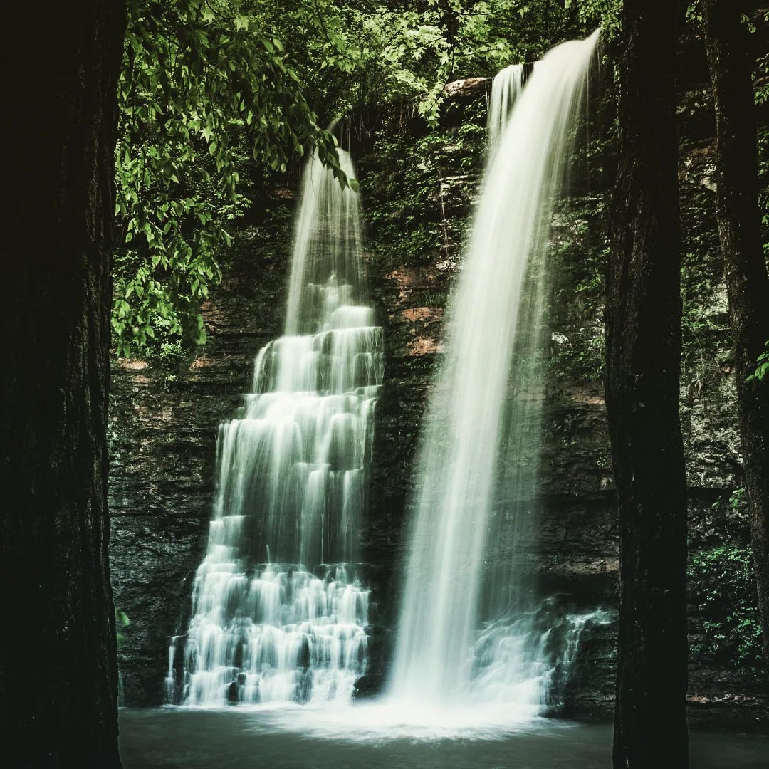 Two small but long, elegant waterfalls are parallel to one another. Their waters pour into the same pool surrounded by lush tall trees deep in the Arkansas Ozarks. 