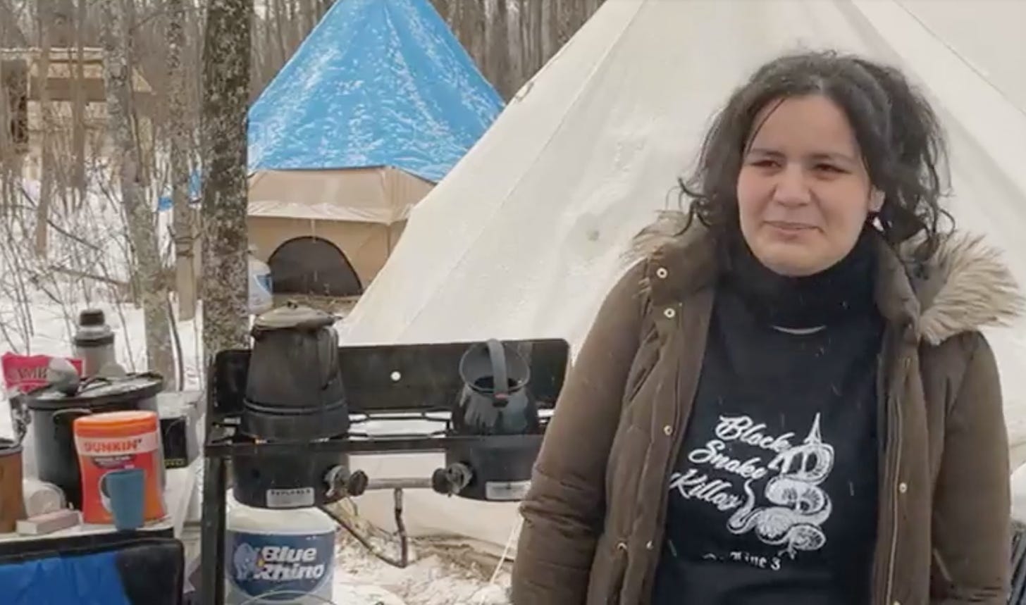 A woman in a black sweater and brown coat, stands in front of a campsite with a gas stove and two tents in the snow