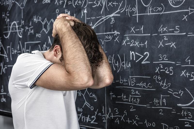 A person standing in front of a blackboard covered in mathematical equations. The person has bowed their head and clasped their hands behind their head, hiding their face in their elbows.