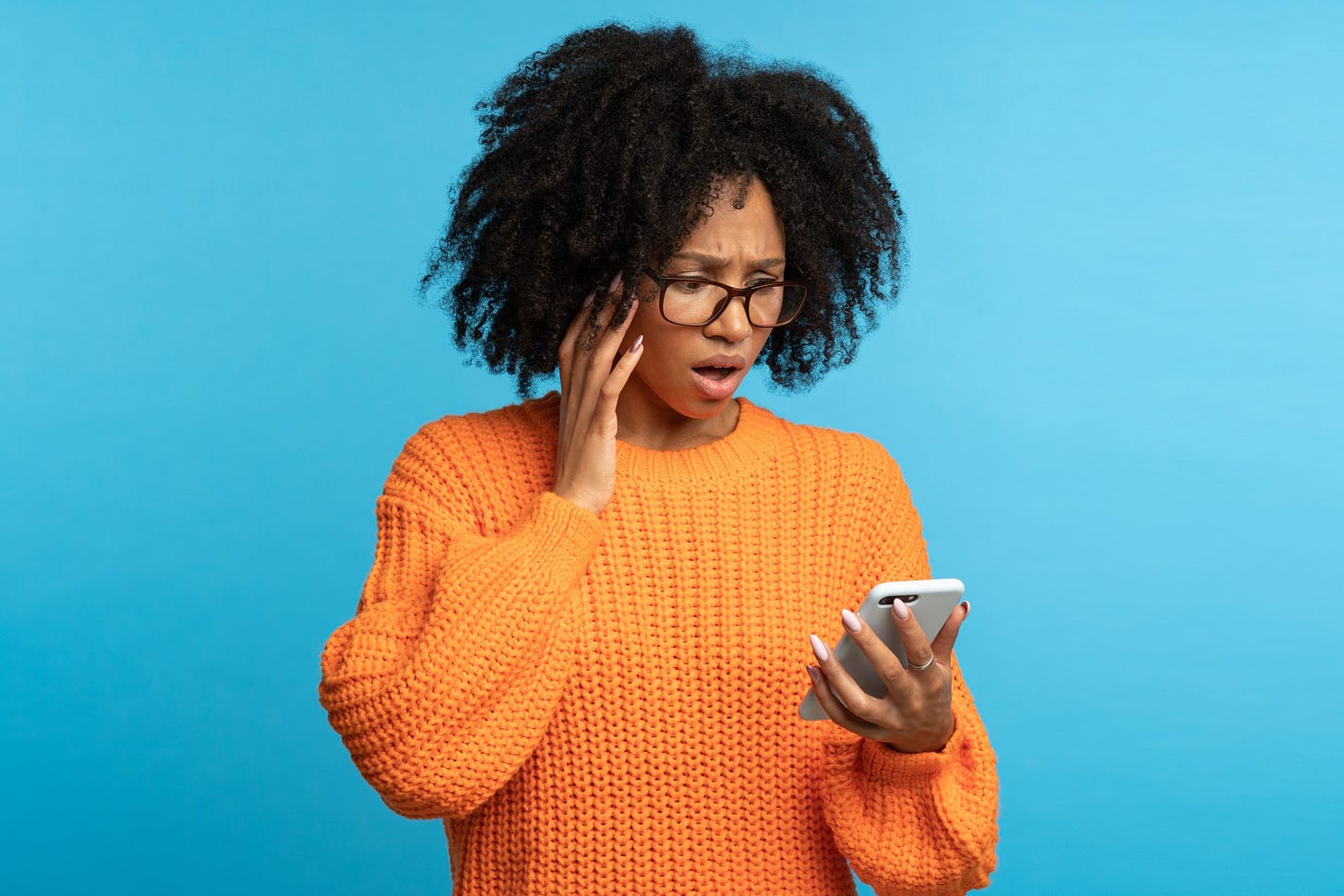 A woman in an orange jumper and curly black hair stares down in shock at her cell phone.